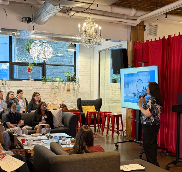 Karen Wonders presenting a "Be Curious" slide at the INWED Toronto 2026 workshop, microphone in hand, with a seated audience.